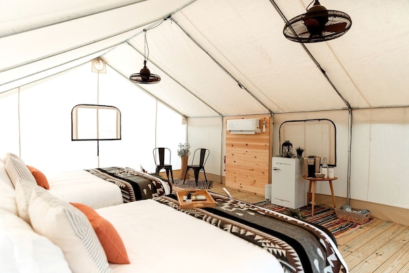 A tented accommodation with a bed, chairs, a small table, a mirror, and a refrigerator.