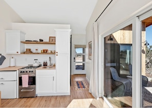 A compact kitchen with white cabinets, a stove, and a sink. There is a dining area with a table and chairs, and a large sliding glass door leading to an outdoor space.