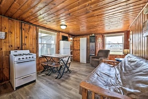Ein Ferienhausinnenraum mit Holzbalkendecke, einer Küchenzeile mit Herd, einem Esstisch, einer Couch und Blick durch ein Fenster.