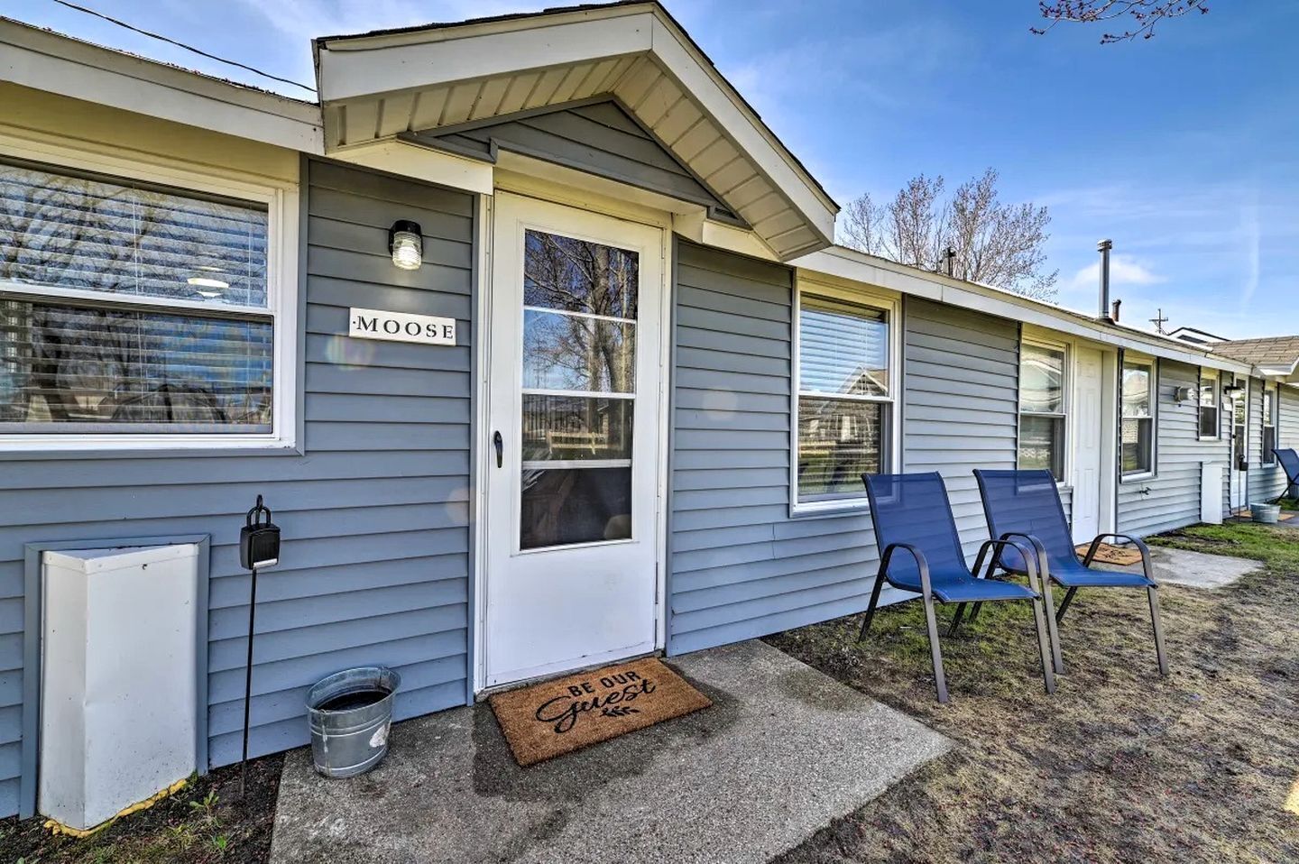 A row of mobile homes with a sign reading 'MOOSE' on the first one, a doormat with 'BE OUR Guests' on the front door, and two blue chairs outside.