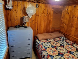 Een slaapkamer met een houten wand, een bed met een geborduurd quilt, een dressoir, een ventilator en een klok.