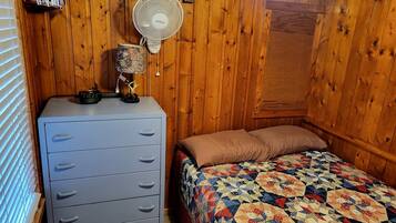 A bedroom with a wooden wall, a bed with a patterned quilt, a dresser, a fan, and a clock.