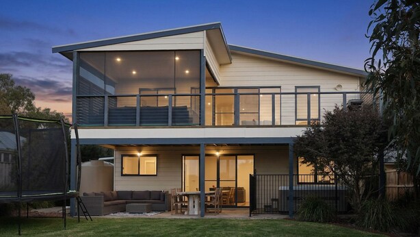 Una casa de dos pisos con un balcón, un patio con mobiliario exterior y un gran ventanal.
