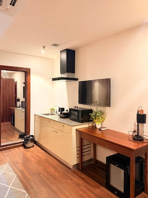 Una cocina funcional con microondas, una mesa pequeña, un televisor montado en la pared y una puerta visible que da a otra habitación.