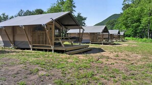 A row of wooden cabins with elevated platforms and attached awnings, set in a grassy area with trees in the background. - Safari Tent Retreat with Riverfront Deck and Nature Stay in Blacksburg, Virginia (Blacksburg)