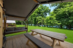 Un patio al aire libre con una mesa y bancos de madera, un espacio de estar cubierto y vistas a un lago rodeado de árboles.