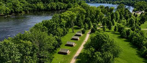 Ein Campingplatz mit Zelten, die an einem Feldweg entlang aufgereiht sind, umgeben von üppiger Vegetation und einem See.