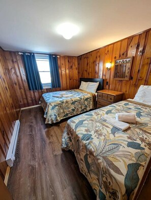 A room with wooden walls and floors, two beds with floral bedding, a nightstand, and a wall-mounted light fixture. - Elegant Oceanfront Cottages with Private Beach Access in North Truro, Massachusetts (North Truro)