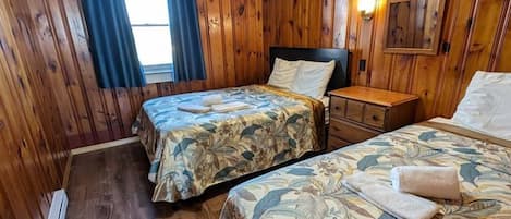A room with wooden walls and floors, two beds with floral bedding, a nightstand, and a wall-mounted light fixture.
