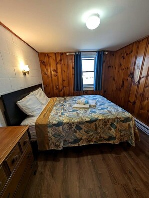 Une pièce avec un lit, des murs en bois, une fenêtre avec des rideaux et un luminaire fixé au mur.