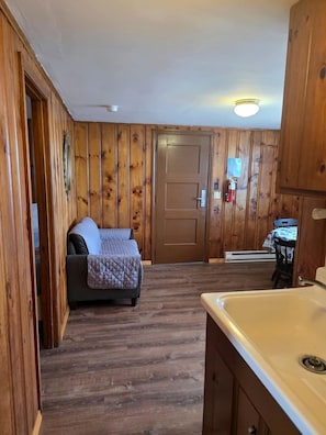 Une pièce aux murs lambrissés, un lit avec une couverture, un lavabo et une porte donnant sur une autre pièce.