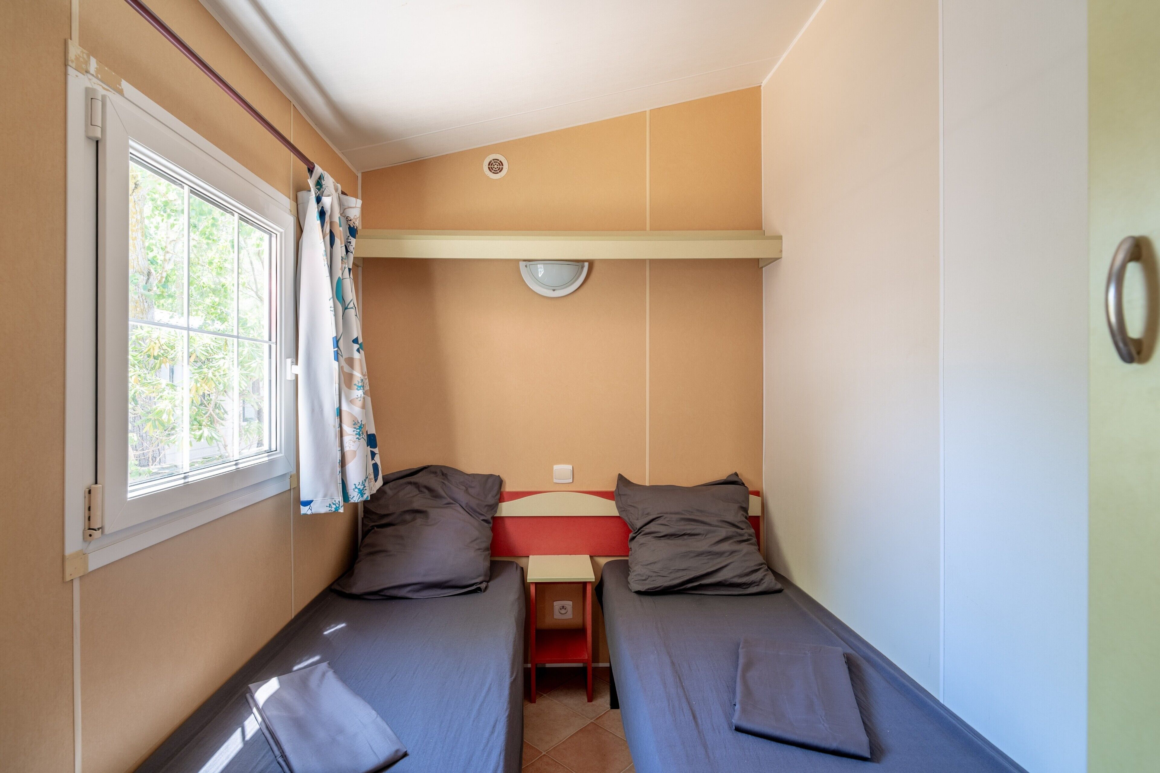 Une petite pièce, de taille modeste, comprenant deux lits simples, une fenêtre avec des rideaux, une petite table et un luminaire fixé au mur.