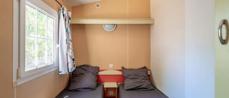 A small, compact room with two single beds, a window with curtains, a small table, and a wall-mounted light fixture.