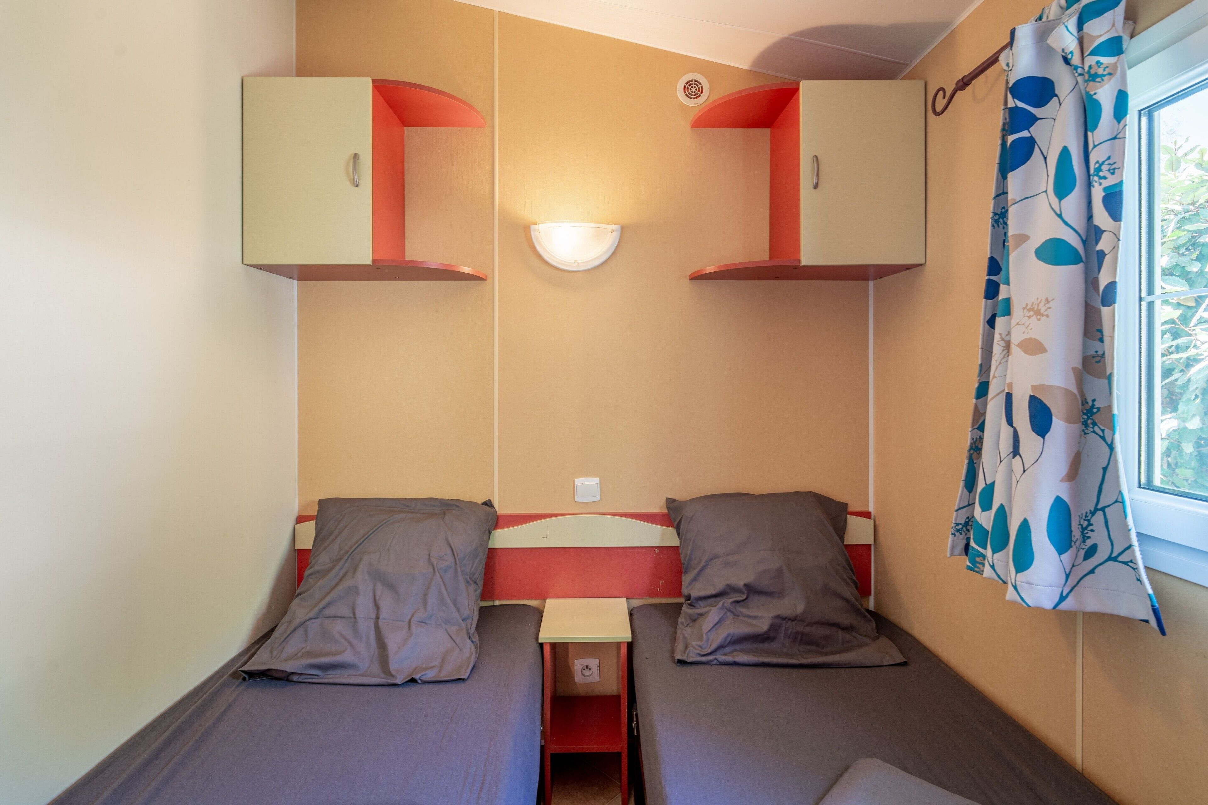 A small, compact room with two single beds, a wall-mounted light, and a window with curtains.