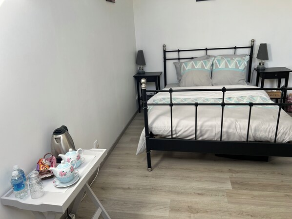 A neatly arranged bedroom with a bed, bedside tables, a small table with tea set, and a kettle.