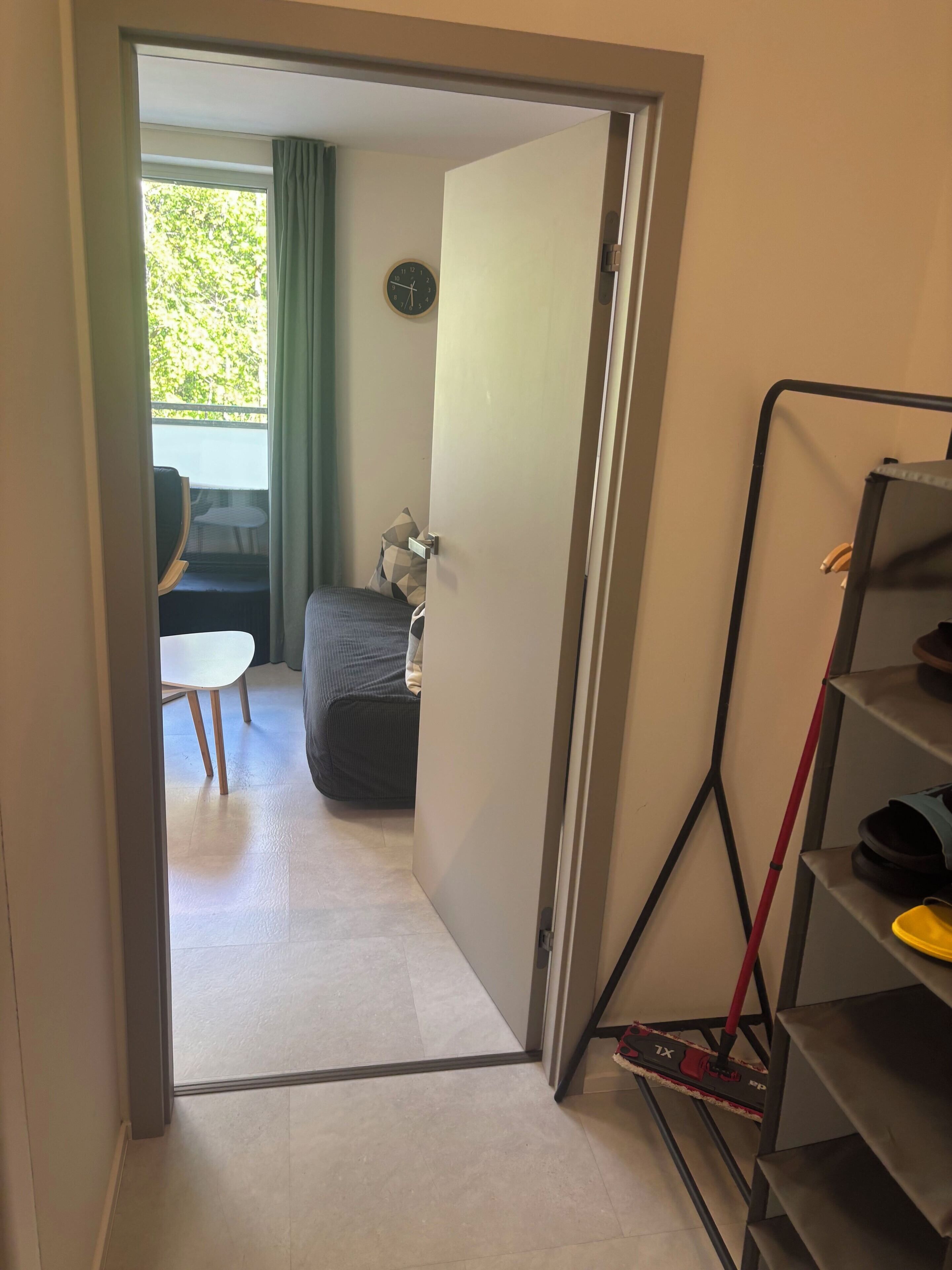 A room with a bed, a small table, a chair, a clock on the wall, and a door leading to another room.