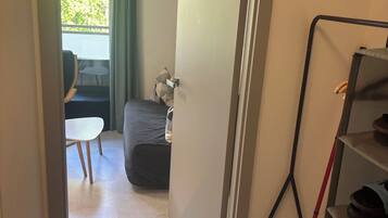 A room with a bed, a small table, a chair, a clock on the wall, and a door leading to another room.