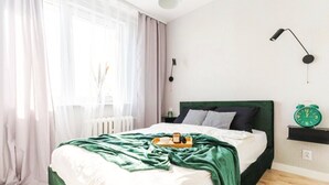 A neatly made bed with a green headboard, a bedside table with a lamp, and a green alarm clock on a shelf.