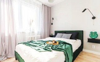 Una cama bien hecha con cabecera verde, una mesita de noche con lámpara y un despertador verde en una estantería.
