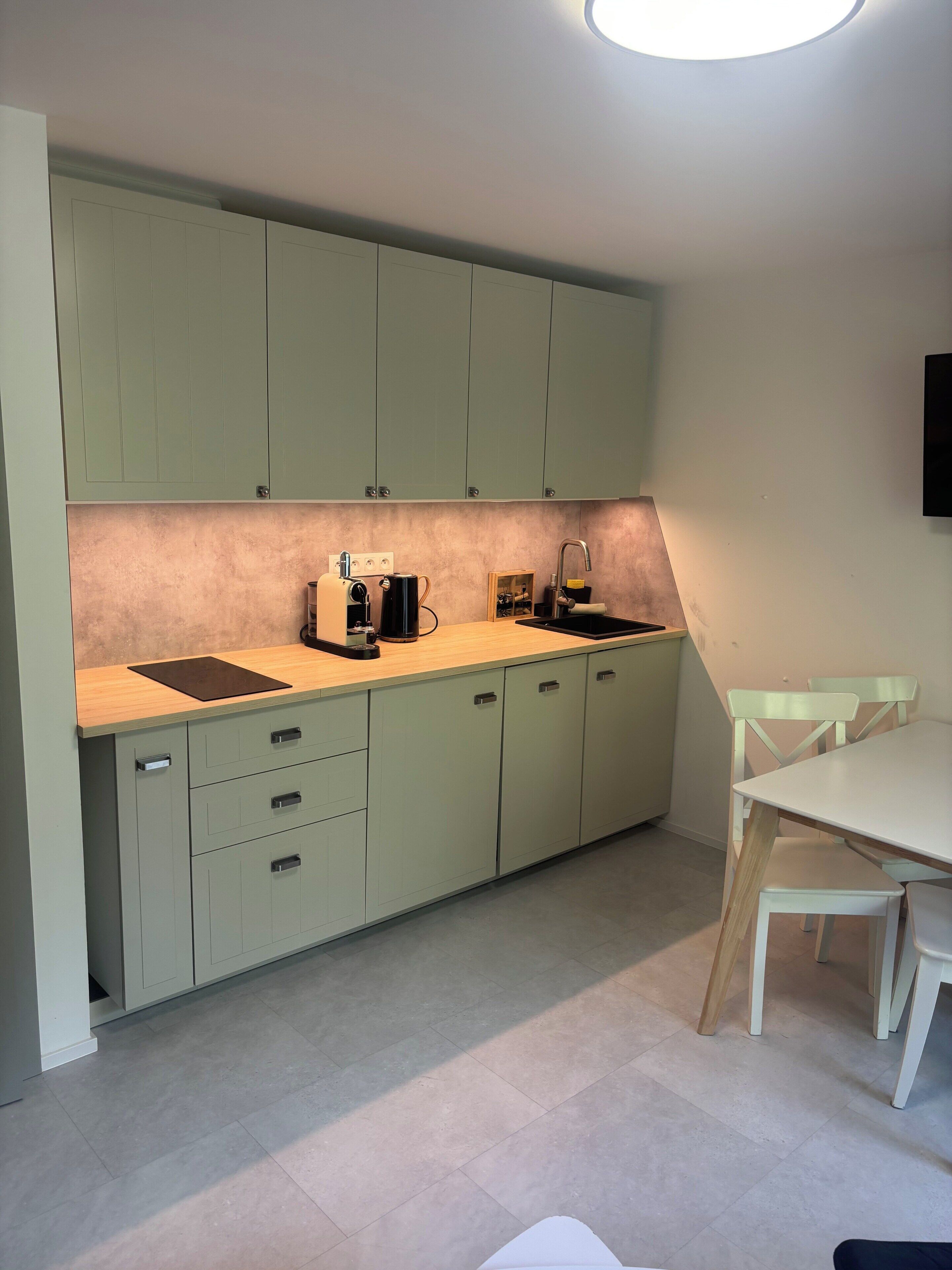A modern kitchen with light green cabinets, a wooden countertop, a sink, and a dining area with white chairs.