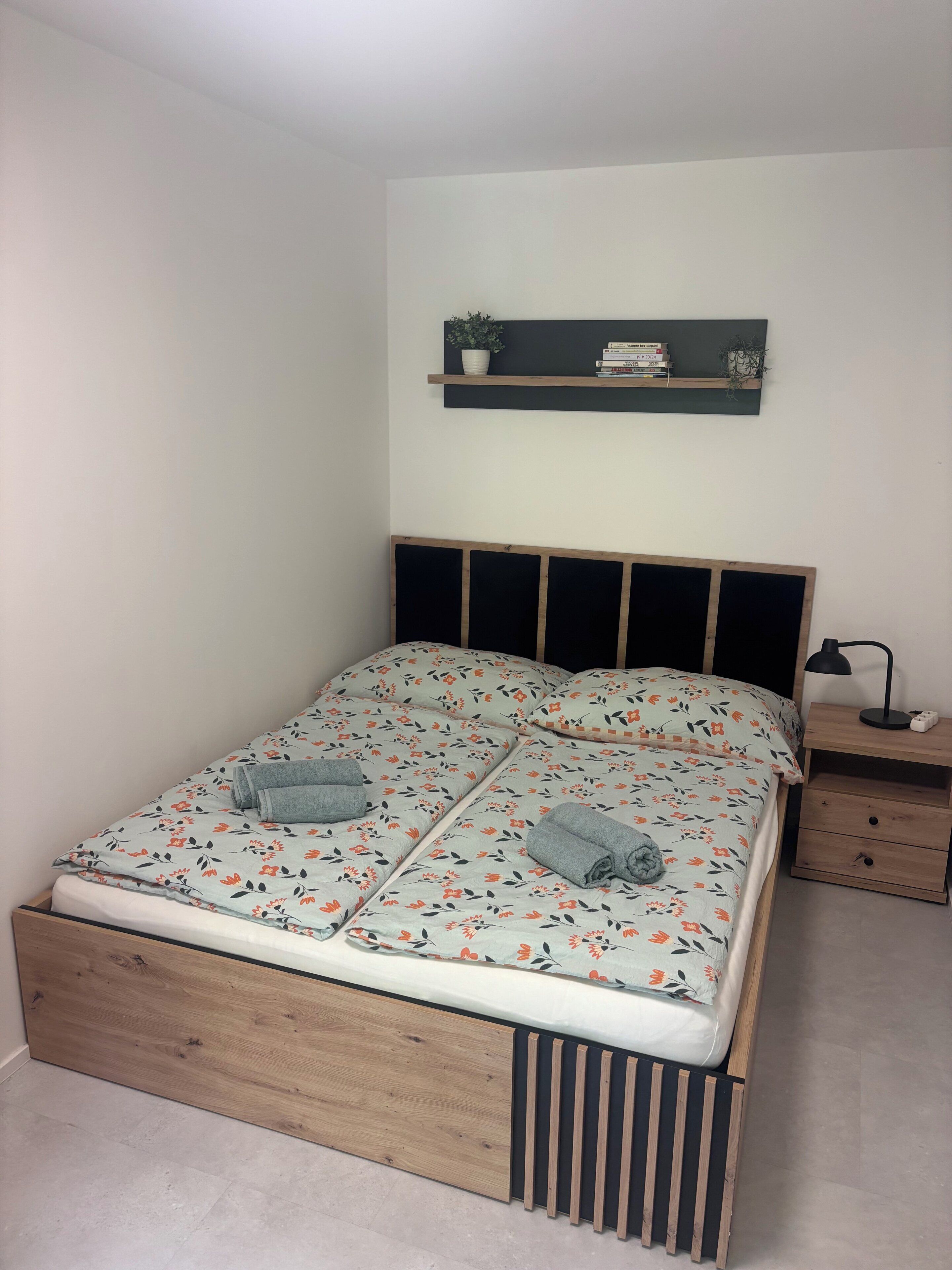 A bed with a floral bedspread, a bedside table with a lamp, and a shelf above the bed.