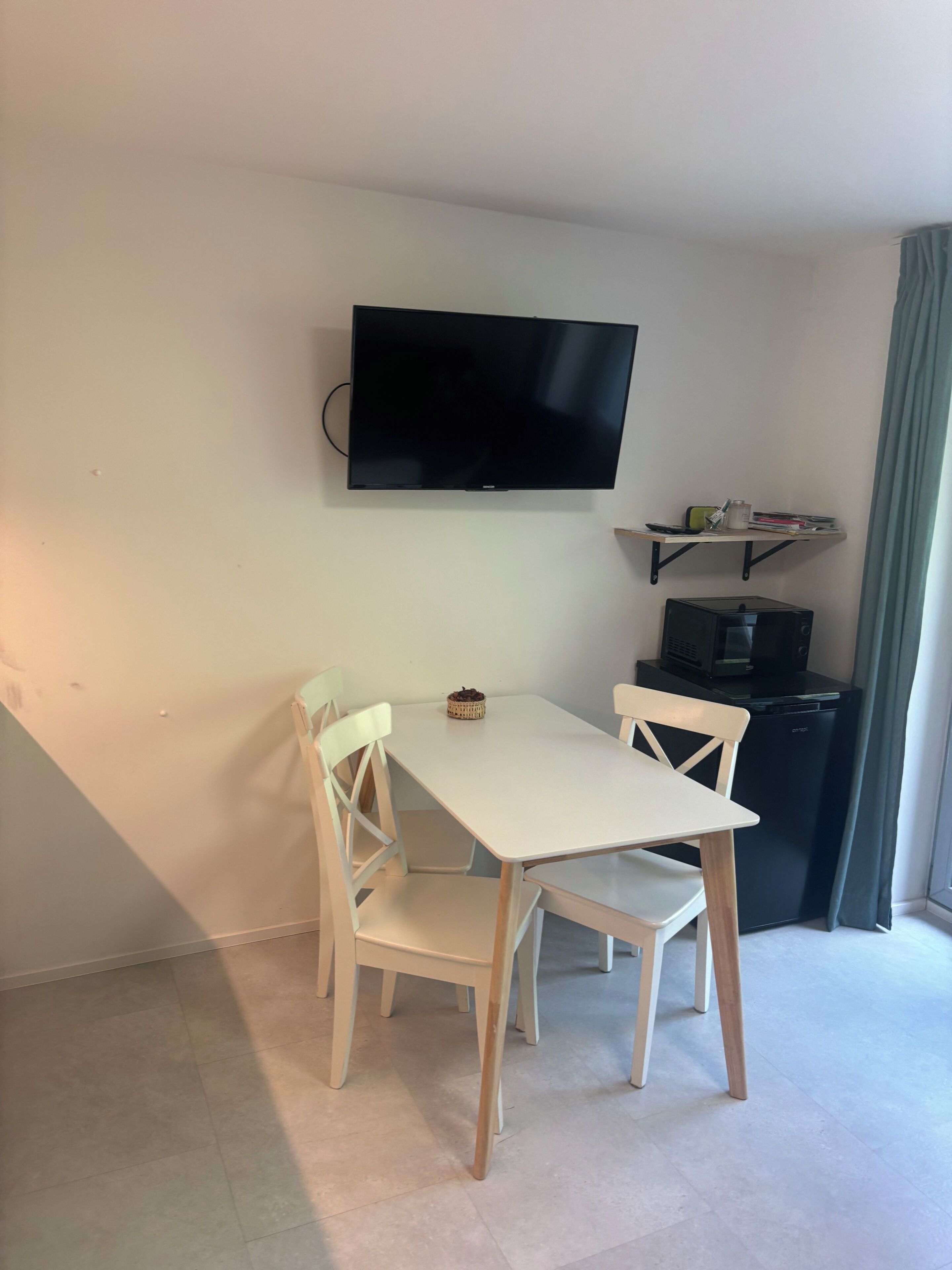 A small dining area with a table and chairs, a flat-screen TV mounted on the wall, a small refrigerator, and a shelf with books and a cup.