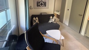 A modern hotel room with a bed, a seating area with a chair and table, a large window, and a camera artwork on the wall.