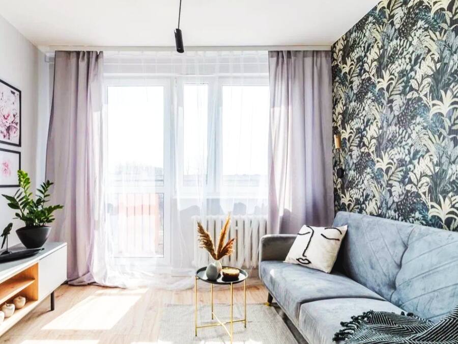 A modern living room with a grey sofa, a black coffee table, a patterned wall, and a large window with sheer curtains.