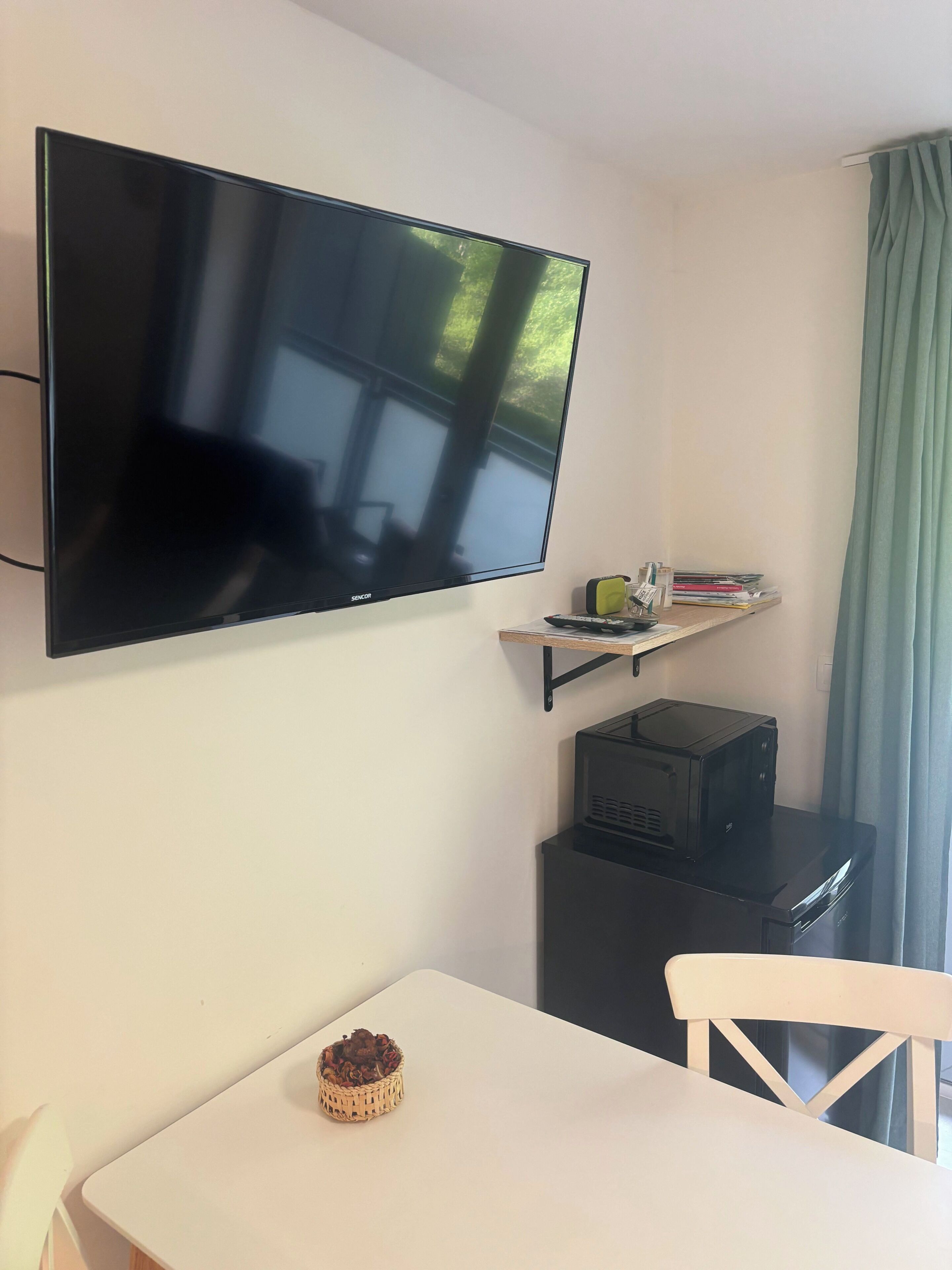 A room with a flat-screen TV mounted on the wall, a small table with a basket, a chair, and a printer on a desk.