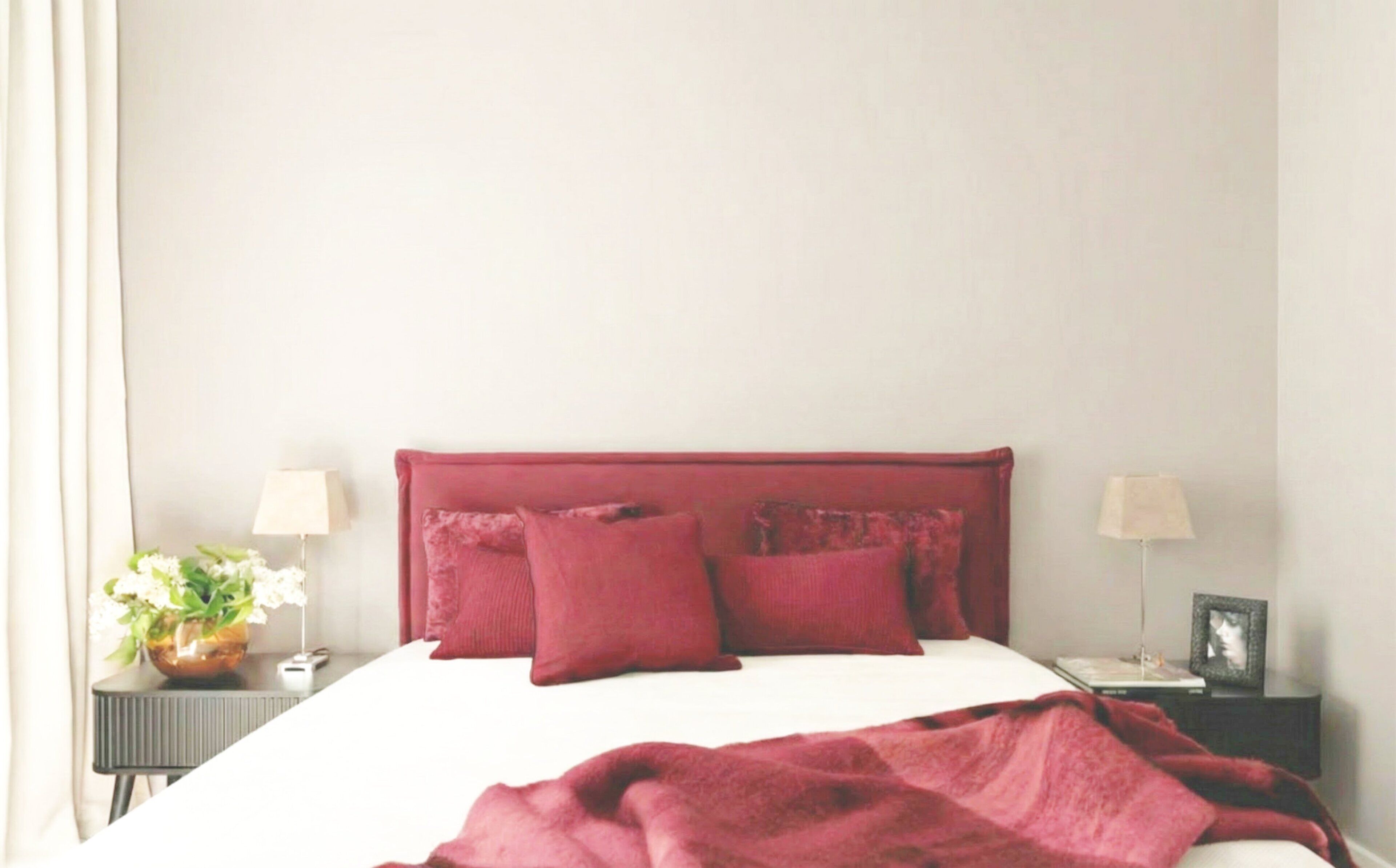 A neatly made bed with a red headboard, two bedside tables with lamps, and a vase with flowers on the left side.