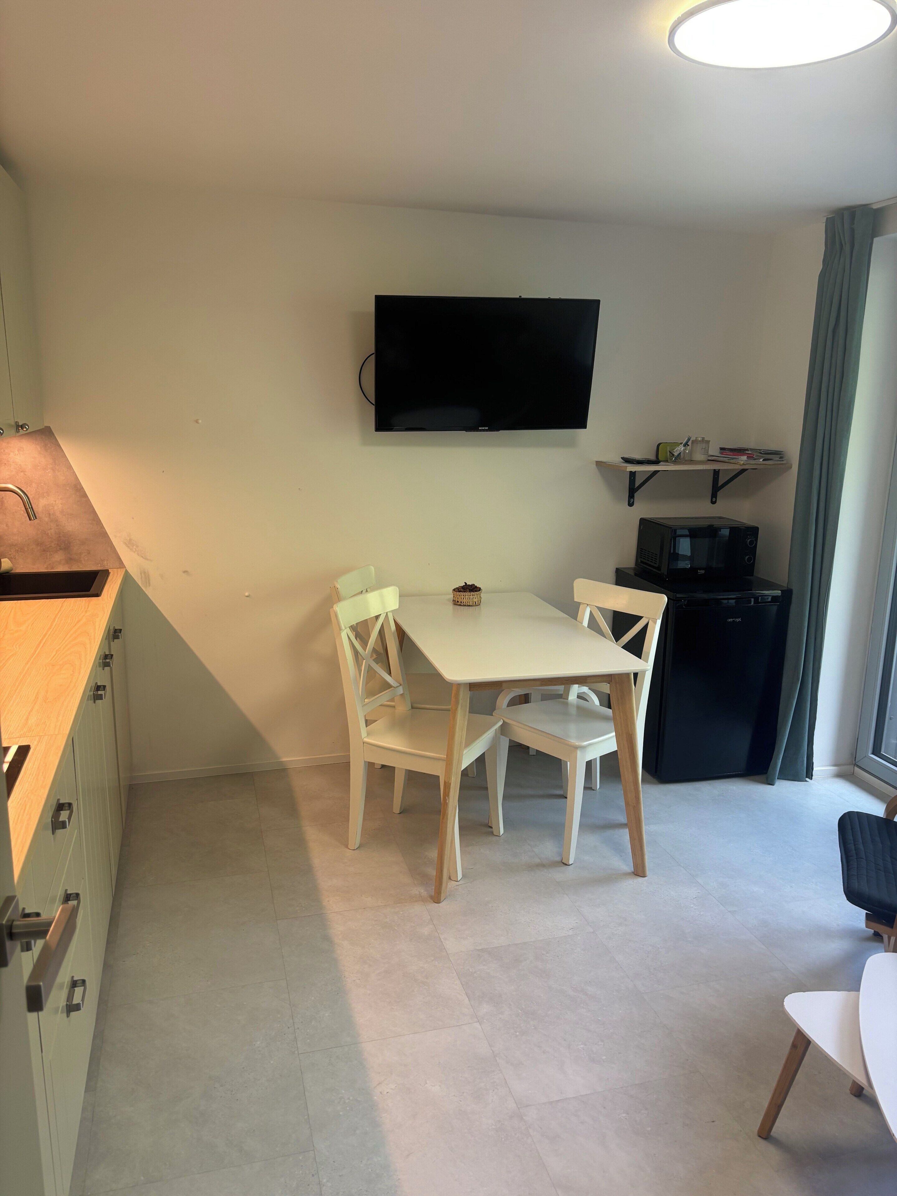 A compact living space with a kitchenette, a dining table with chairs, a flat-screen TV mounted on the wall, a microwave, and a small refrigerator.