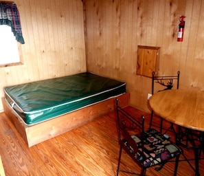 A room with a wooden floor, a green mattress, a small table with chairs, and a fire extinguisher on the wall.