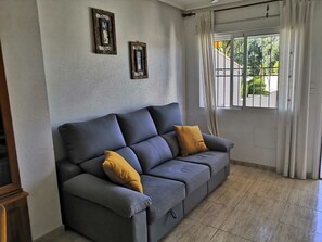 Ein graues Sofa mit zwei Kissen, ein Fenster mit Vorhängen und zwei Bilderrahmen an der Wand.