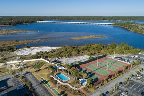 Luftaufnahme eines Küsteneigentums mit Tennisplatz, Schwimmbad und einem großen Gewässer mit Brücke im Hintergrund.