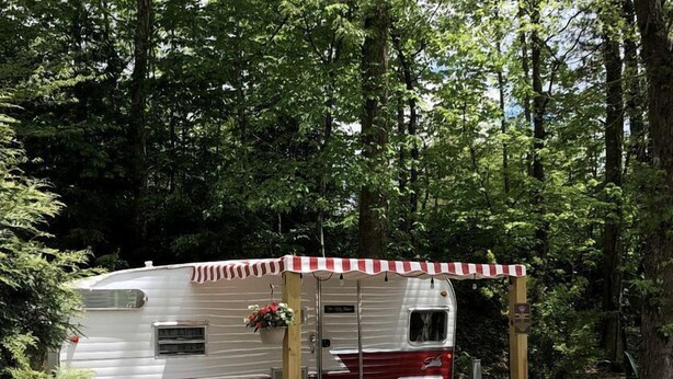 Un véhicule récréatif doté d’un auvent rayé rouge et blanc, entouré d’une zone de gravier où sont disposées des chaises Adirondack rouges.
