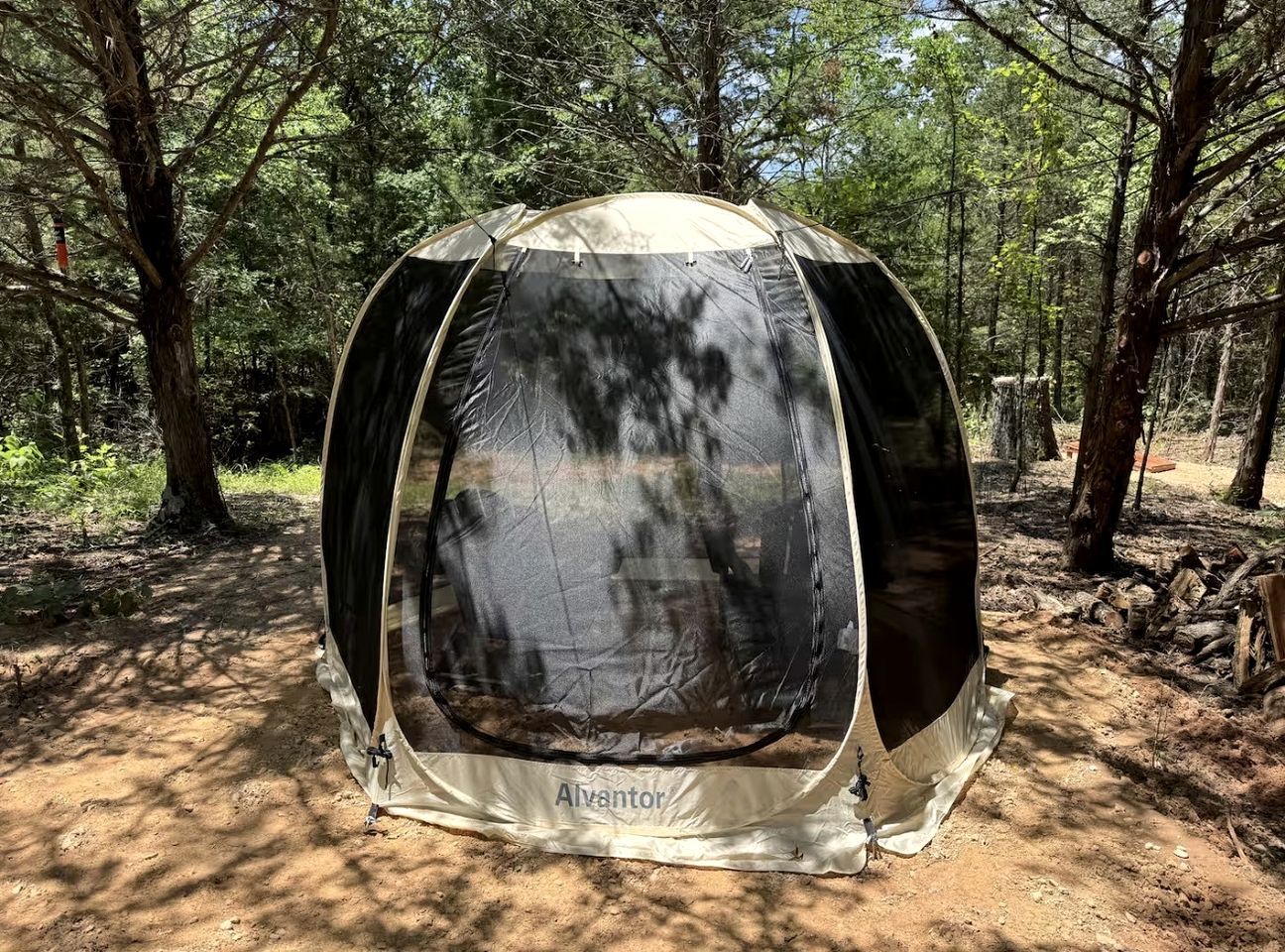Une tente en forme de dôme, dotée d’un toit en toile, portant l’inscription « Alvantor », installée dans une zone boisée avec des arbres et un sol en terre.