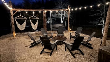 Un espace extérieur aménagé avec des chaises et un foyer, entouré d’une pergola en bois illuminée de guirlandes lumineuses.