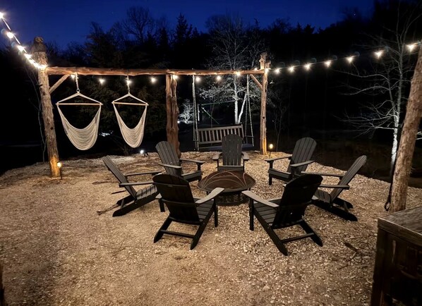 An outdoor seating area with chairs and a fire pit, surrounded by a wooden pergola with string lights.