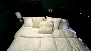 A bed with a white duvet and several pillows, a bedside lamp, and a small picture frame on the wall. - Luxurious Remodeled Airstream with Wraparound Deck and Spa near Honey Grove, Texas (Honey Grove)