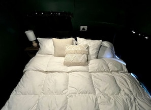 Ein Bett mit weißer Bettdecke und mehreren Kissen, einer Nachttischlampe und einem kleinen Bilderrahmen an der Wand.