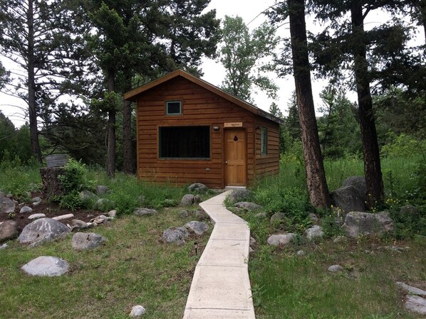 A wooden cabin with a concrete pathway leading to it, surrounded by trees and rocks.