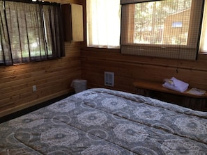Ein Zimmer in einer Holzhütte mit einem Bett, einer Bank, einem Fenster mit Vorhängen und einer Wandleuchte.