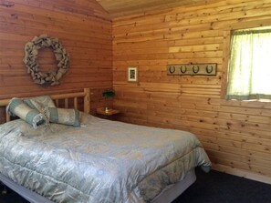 Une pièce de style cabane en bois, avec un lit, une petite table et une lampe, un porte-manteau fixé au mur et une fenêtre avec des rideaux.