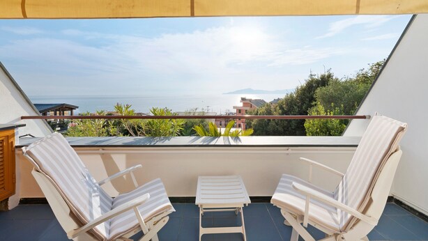 Ein Balkon mit zwei weißen Streifenliegen, einem kleinen Tisch und Blick auf das Meer sowie entfernte Gebäude.