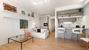 Ein modernes Wohnzimmer mit Glastisch, weißem Sofa und einer Küchenzeile mit Barhocker.