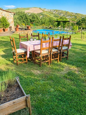 A wooden table with chairs set outdoors, a stone building, and a pool in the background. - An old Tuscan stone farmhouse with a pool and vineyard in the Val d'Orcia (Radicofani)