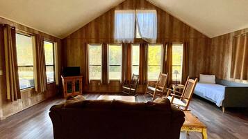 A room with a sloped ceiling, wooden walls, a sofa, two rocking chairs, a bed, and large windows.