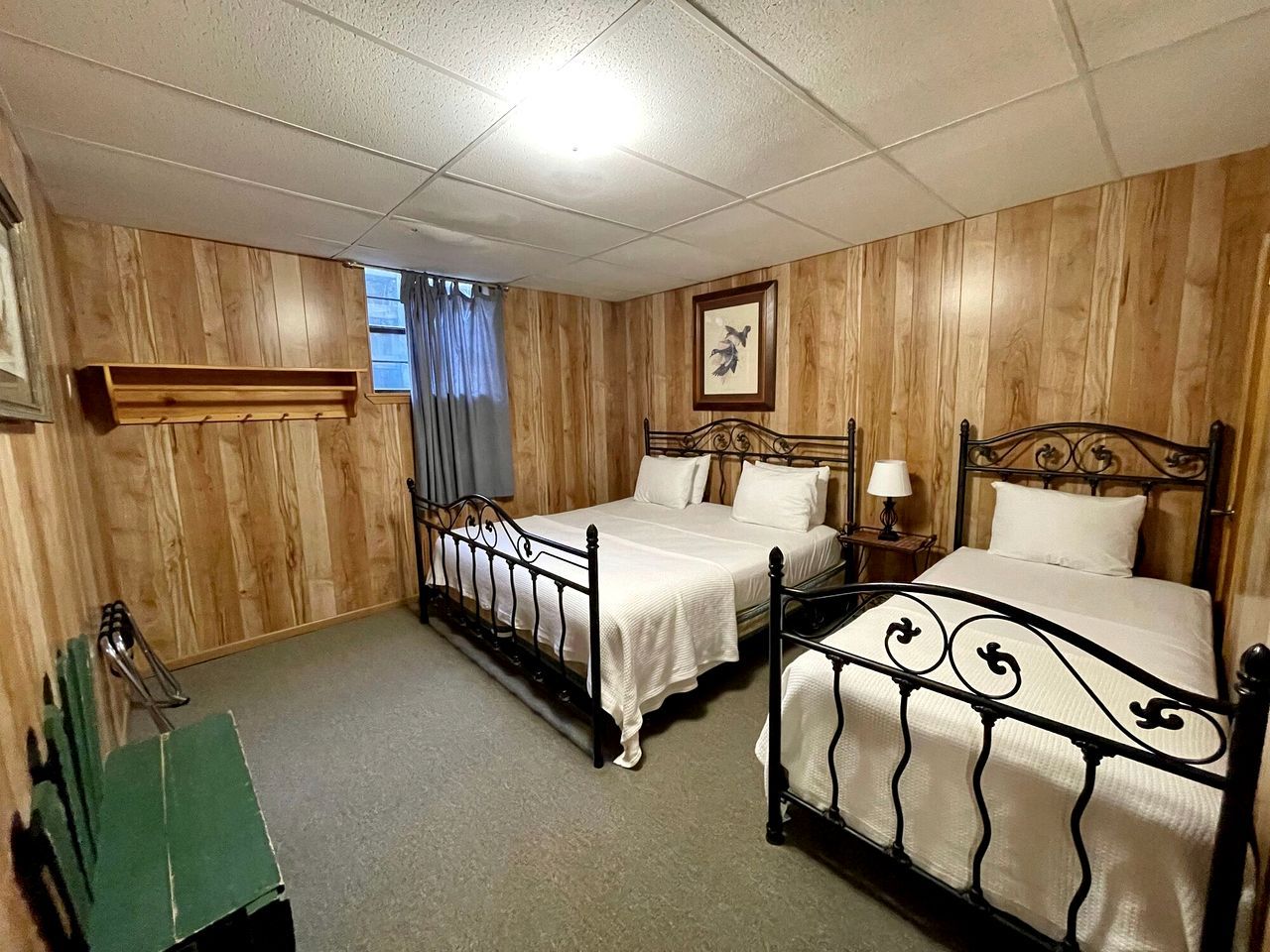 Ein Zimmer mit zwei Betten, Holzvertäfelung, einem mit Vorhängen versehenen Fenster, einem gerahmten Bild an der Wand und einem kleinen Tisch mit einer Lampe.