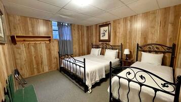Ein Zimmer mit zwei Betten, Holzvertäfelung, einem mit Vorhängen versehenen Fenster, einem gerahmten Bild an der Wand und einem kleinen Tisch mit einer Lampe.