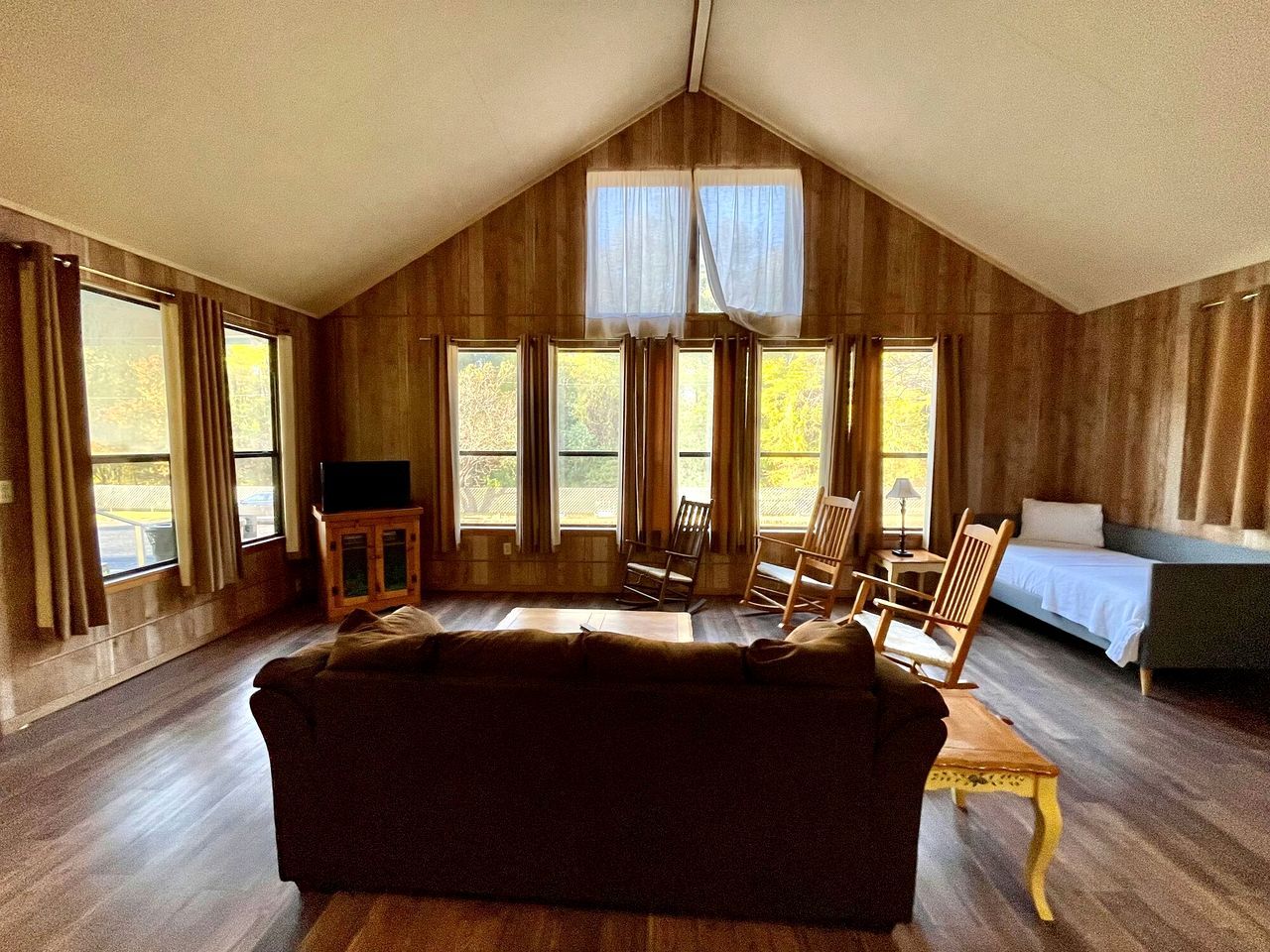 Ein Zimmer mit schräge Decke, Holzwänden, einer Couch, zwei Schaukelstühlen, einem Bett und großen Fenstern.